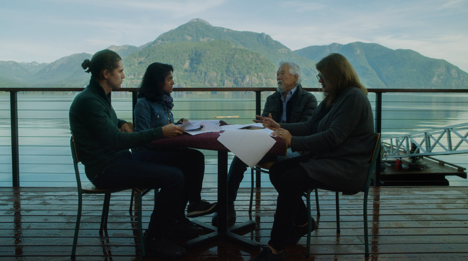 Four people sit around a table on a wooden deck overlooking a calm lake with forested mountains in the background. They are engaged in a serious discussion, holding papers and notes. The setting is peaceful and scenic, with natural light and reflections on the water enhancing the contemplative atmosphere.