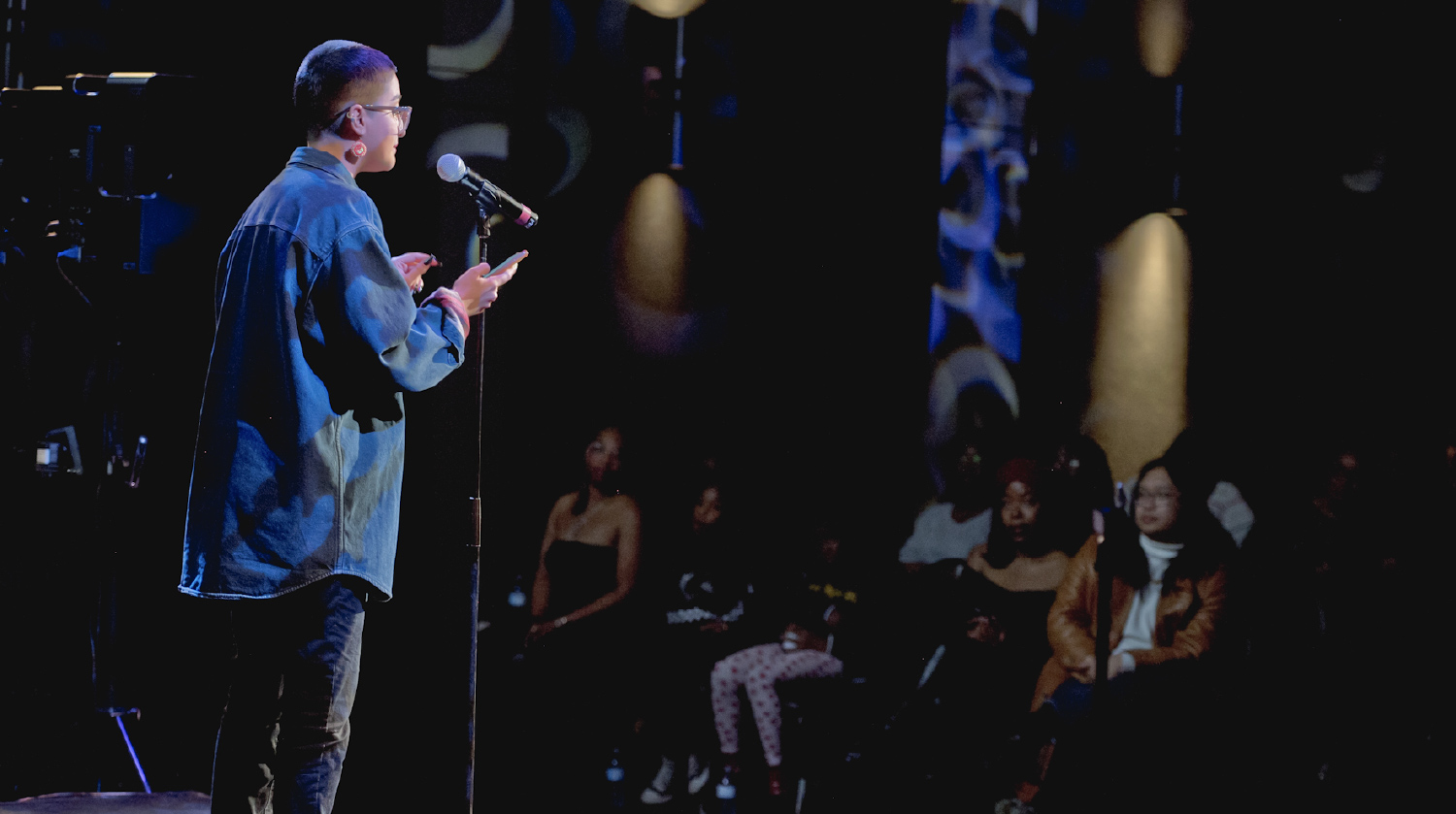 A performer stands on stage under soft lighting, speaking into a microphone. They wear glasses, hoop earrings, and a loose denim jacket, holding notes while addressing the audience. In the dimly lit background, several audience members sit attentively, some with focused expressions as they watch the performance.