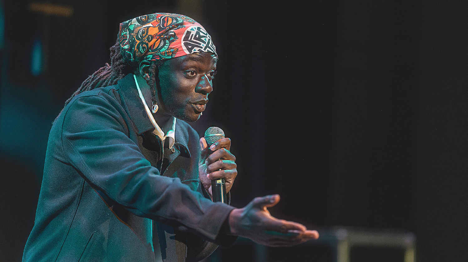 A male performer passionately speaks into a microphone on stage, extending one hand expressively. He wears a colourful patterned headwrap, earrings, and a dark coat. The background is dimly lit, keeping the focus on his animated expression and gesture.