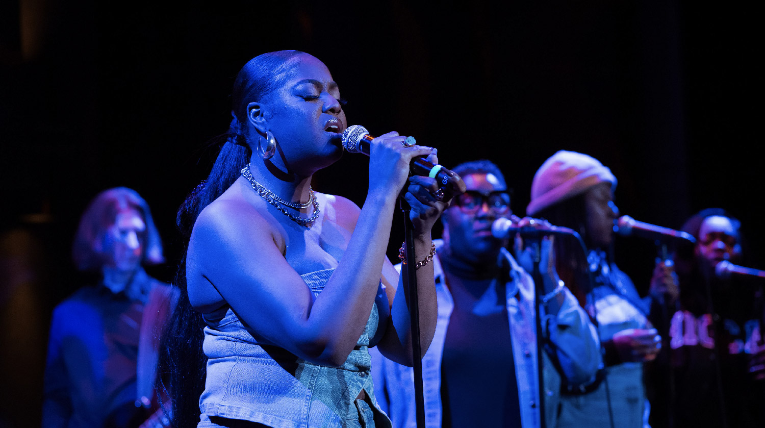 A vocalist passionately singing into a microphone on stage, wearing a denim corset top and jewelry. Behind her, backup singers and a guitarist provide accompaniment, all illuminated by stage lighting.
