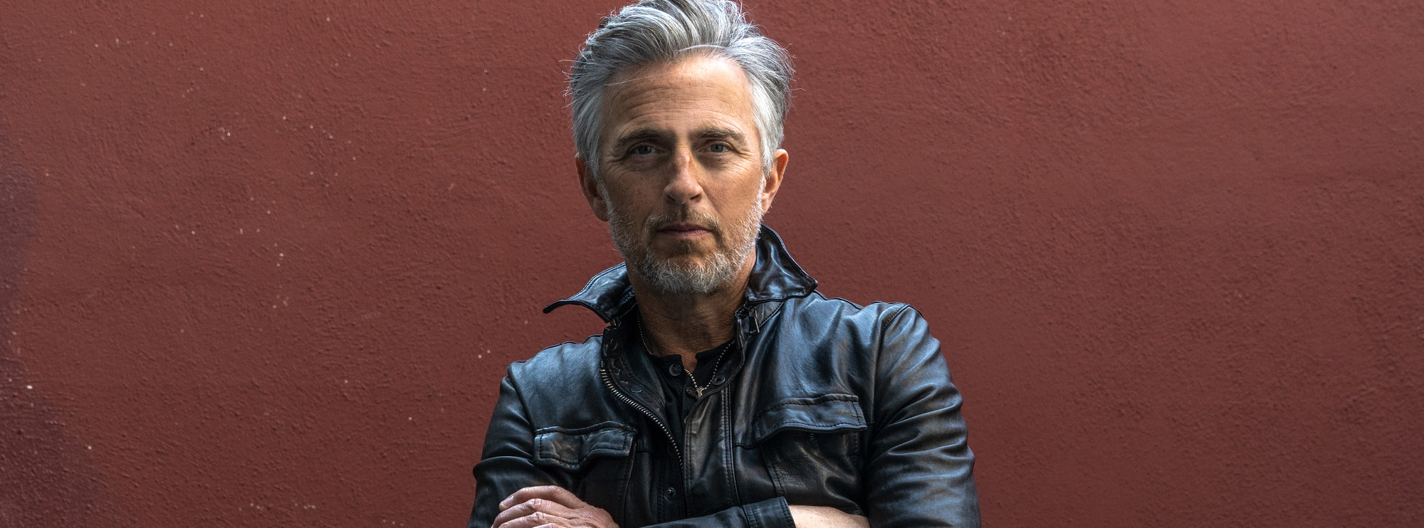 Image of Colin James with his arms crossed atop his body, standing before a dark red wall. He has warm-toned skin, grey hair and a grey beard. He wears a black leather jacket and looks straight ahead.