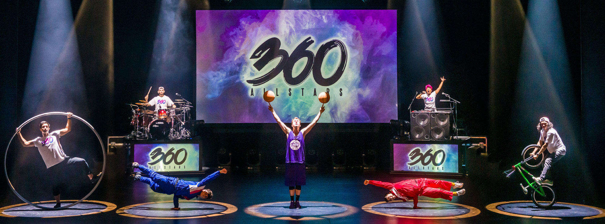 Image of the 360 All Stars perfoming with their logo projected on a screen centre stage. To the left, a man plays the drums atop a platform that reads "360 All Stars" in green, purple and blue. He wears a white shirt with a backwards baseball cap. Standing opposite him on another platform, a woman plays music from a sound system with two large speakers. She speaks into the microphone, and raises her left arm in the air. To the left, a man wearing blue ensemble balances his body on one arm, which is propelled sideways in the air. The man opposite him performs the same stunt, but wears a red suit. In the centre of the image, a man in a purple tank top spins two basketballs on his index fingers. The performs are all illuminated on stage by a colourful spotlight.