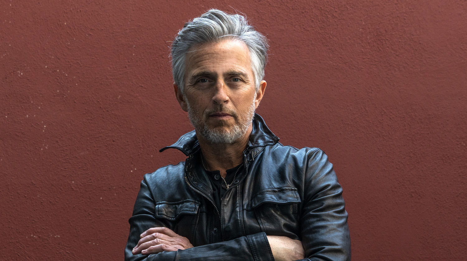 Image of Colin James with his arms crossed atop his body, standing before a dark red wall. He has warm-toned skin, grey hair and a grey beard. He wears a black leather jacket and looks straight ahead.