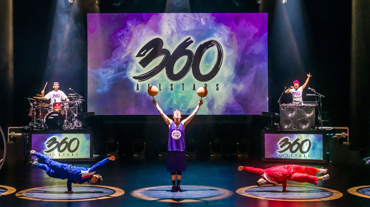 Image of the 360 All Stars perfoming with their logo projected on a screen centre stage. To the left, a man plays the drums atop a platform that reads "360 All Stars" in green, purple and blue. He wears a white shirt with a backwards baseball cap. Standing opposite him on another platform, a woman plays music from a sound system with two large speakers. She speaks into the microphone, and raises her left arm in the air. To the left, a man wearing blue ensemble balances his body on one arm, which is propelled sideways in the air. The man opposite him performs the same stunt, but wears a red suit. In the centre of the image, a man in a purple tank top spins two basketballs on his index fingers. The performs are all illuminated on stage by a colourful spotlight.
