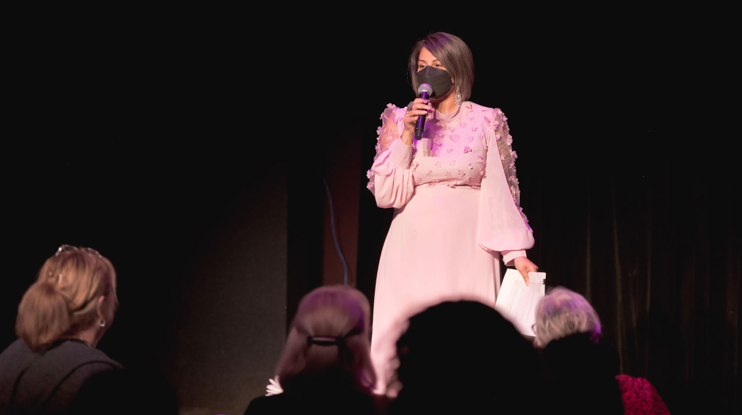 Image of a woman speaking into a microphone before a crowd of seated people. The woman has shoulder length brown hair and medium-toned skin. She wears a black mask and ornate dangling, silver earrings. She is outfitted in a long, light pink dress with sheer flowy sleeves. The top of the dress is embellished with flowers. The room beyond her is dimly lit. 