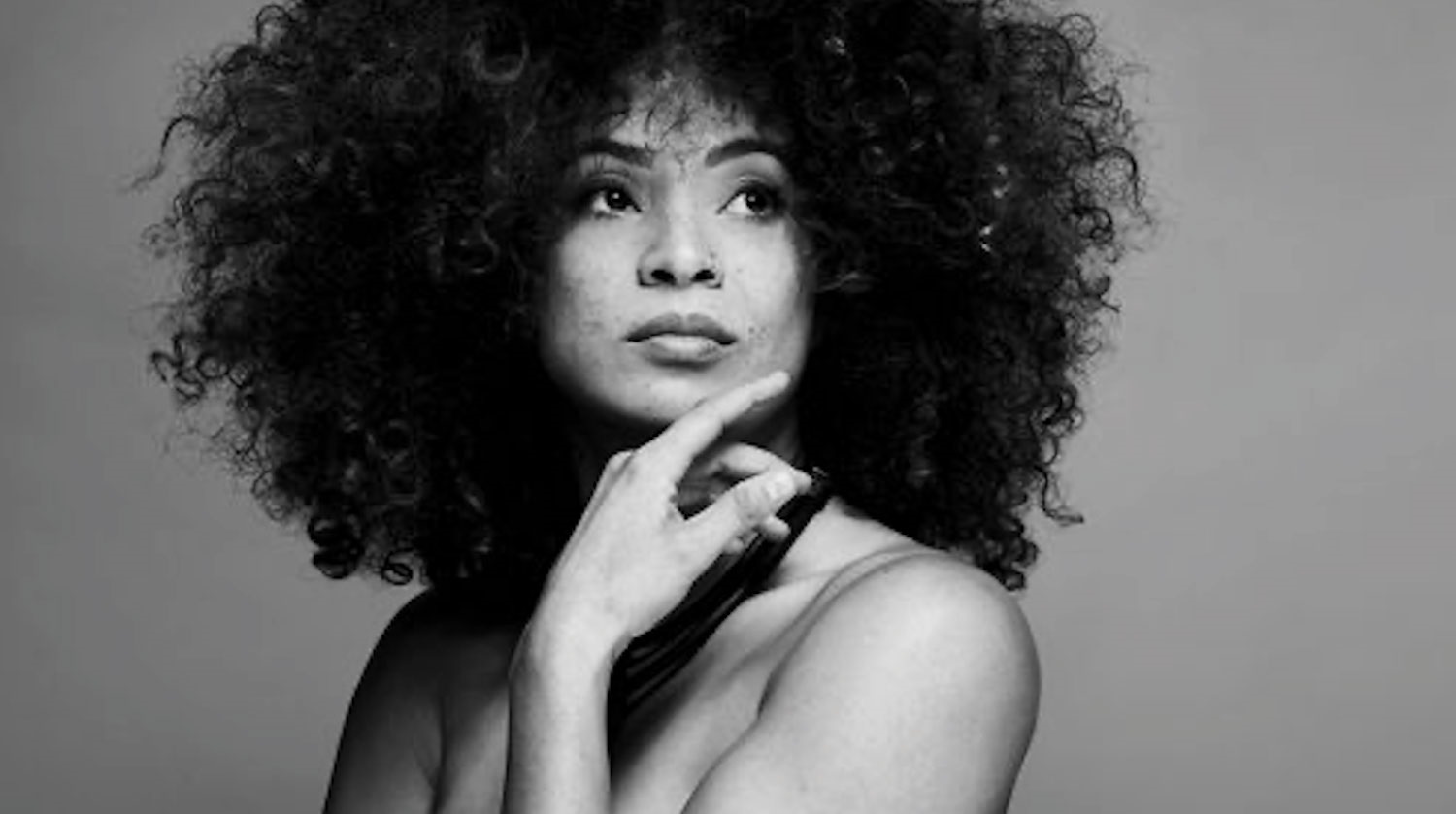 Black and white image of Kandace Springs. She has dark-toned skin, black curly hair, and a nose ring. She is looking out, and has her left hand placed up towards her chin. 