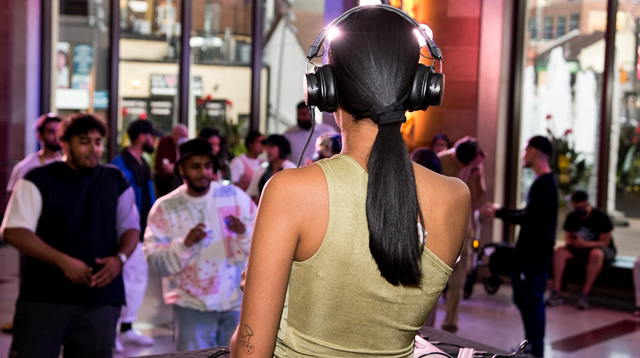 Image of a DJ facing away from the camera toward a crowd of diverse young adults in The Rose Lobby. She has medium skin and is wearing black headphones with her dark hair in a ponytail. She is wearing a champagne-coloured one-shoulder tank top as she spins her DJ equipment on a table in front of her. The crowd is dancing and socializing. The sunlight shines through the large Rose Lobby windows in the background. 