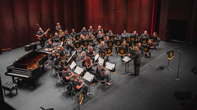 A full orchestra on stage with brass, strings, and a grand piano, music stands adorned with golden logos. 