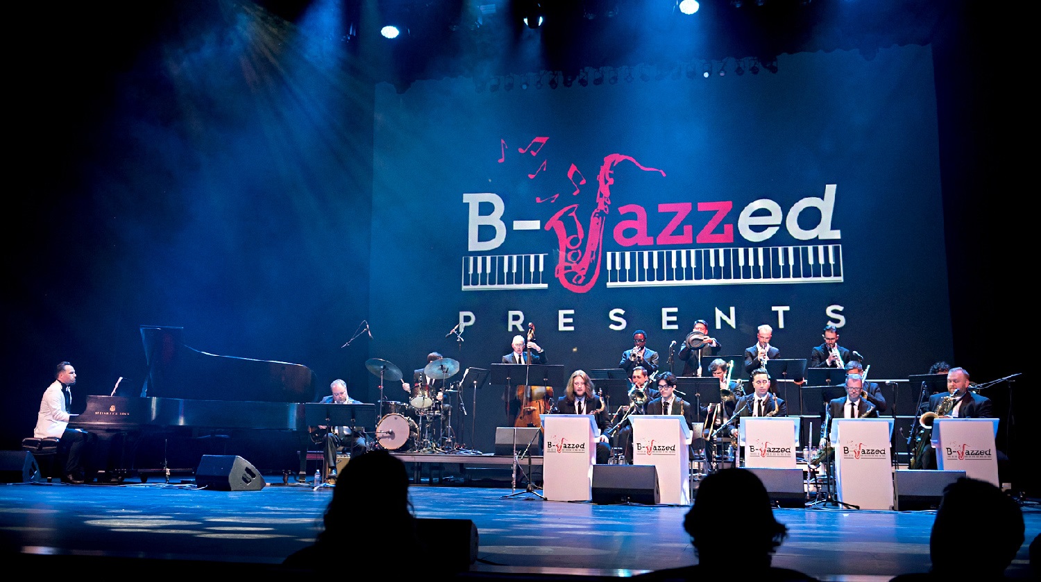 A jazz band performs on stage at the "B-Jazzed Presents" event. The large ensemble includes musicians playing various instruments such as the piano, drums, trumpets, trombones, and saxophones. The stage is bathed in blue lighting, and the backdrop features a large projection of the "B-Jazzed" logo, which includes a stylized saxophone and piano keys.