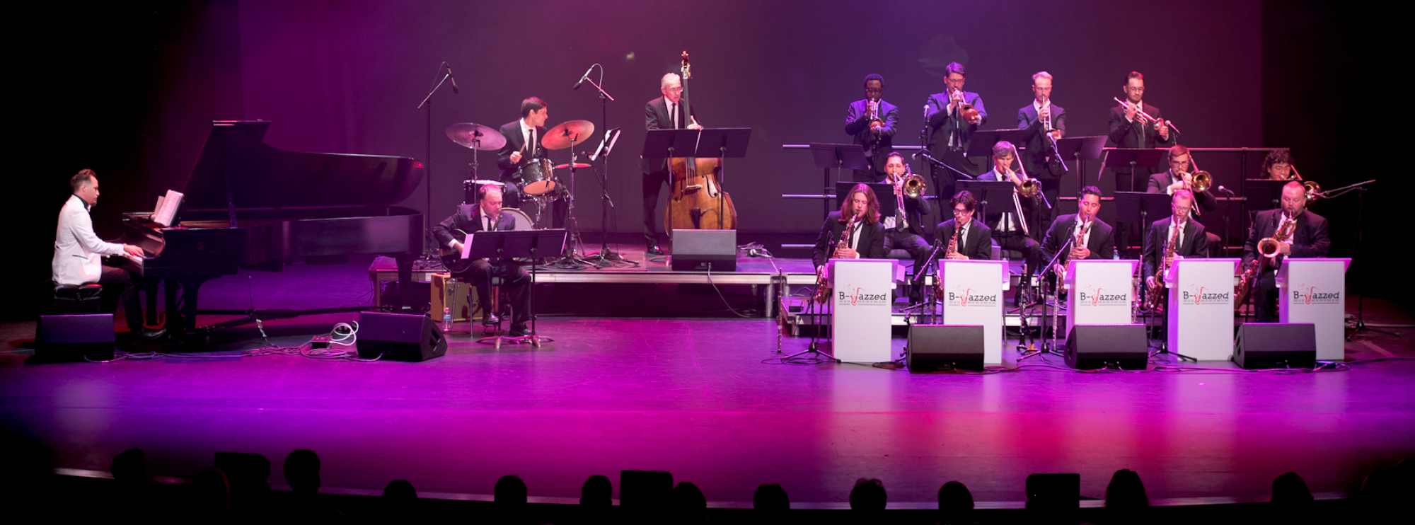 Image of the B-Jazzed band performing on The Rose Mainstage. To the left of the image, several musicians play an array of instruments, including the saxophone, trumpet, and drums. To the right of the image, artist Carmen Spada plays the piano. Behind the performs, a projection reads "B-Jazzed Presents."