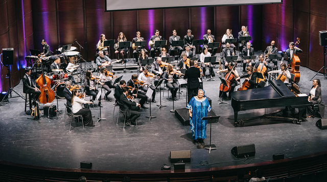 A full orchestra with music stands before them, positioned in concert formation on a stage. The musicians are led by a conductor, with a soloist in blue attire at the foreground. 