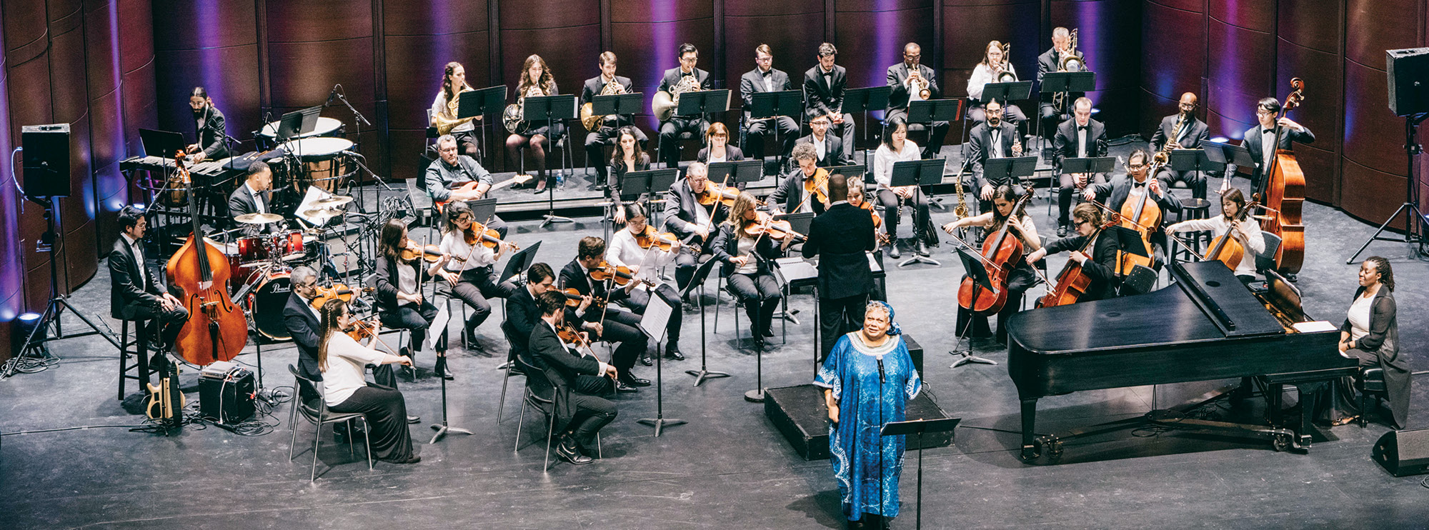 A full orchestra with music stands before them, positioned in concert formation on a stage. The musicians are led by a conductor, with a soloist in blue attire at the foreground. 