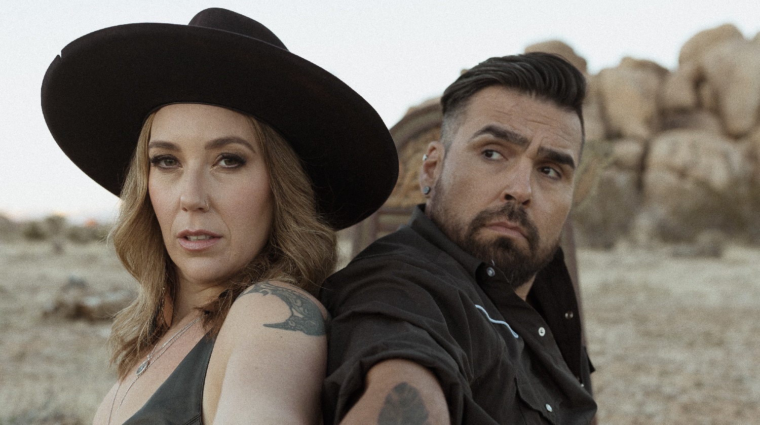 A striking portrait features a woman and a man sitting back-to-back in a rugged outdoor setting, possibly a desert with rocky terrain visible in the background. The woman wears a wide-brimmed black hat and a sleeveless top. The man has a rugged look with short hair, a beard, and is dressed in a dark shirt with white piping.