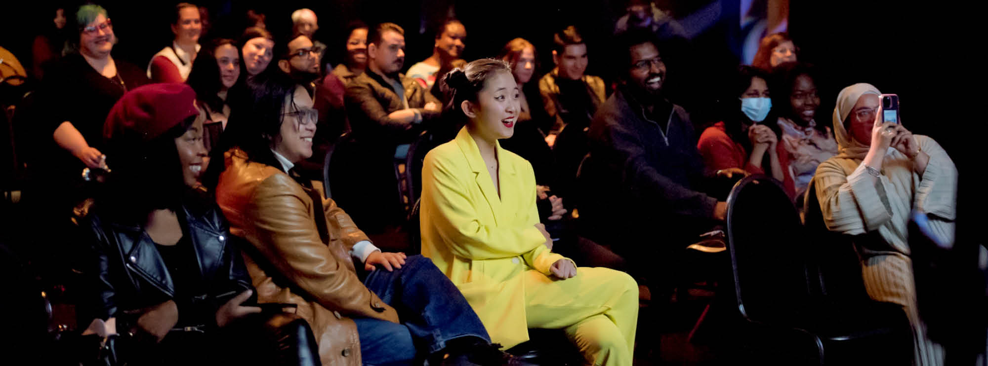 Image of audience members smiling and enjoying a performance in The Rose Studio. One woman holds her phone in front of her to capture the show. Another woman in a yellow matching suit smiles enthusiastically as she observes the performance. 