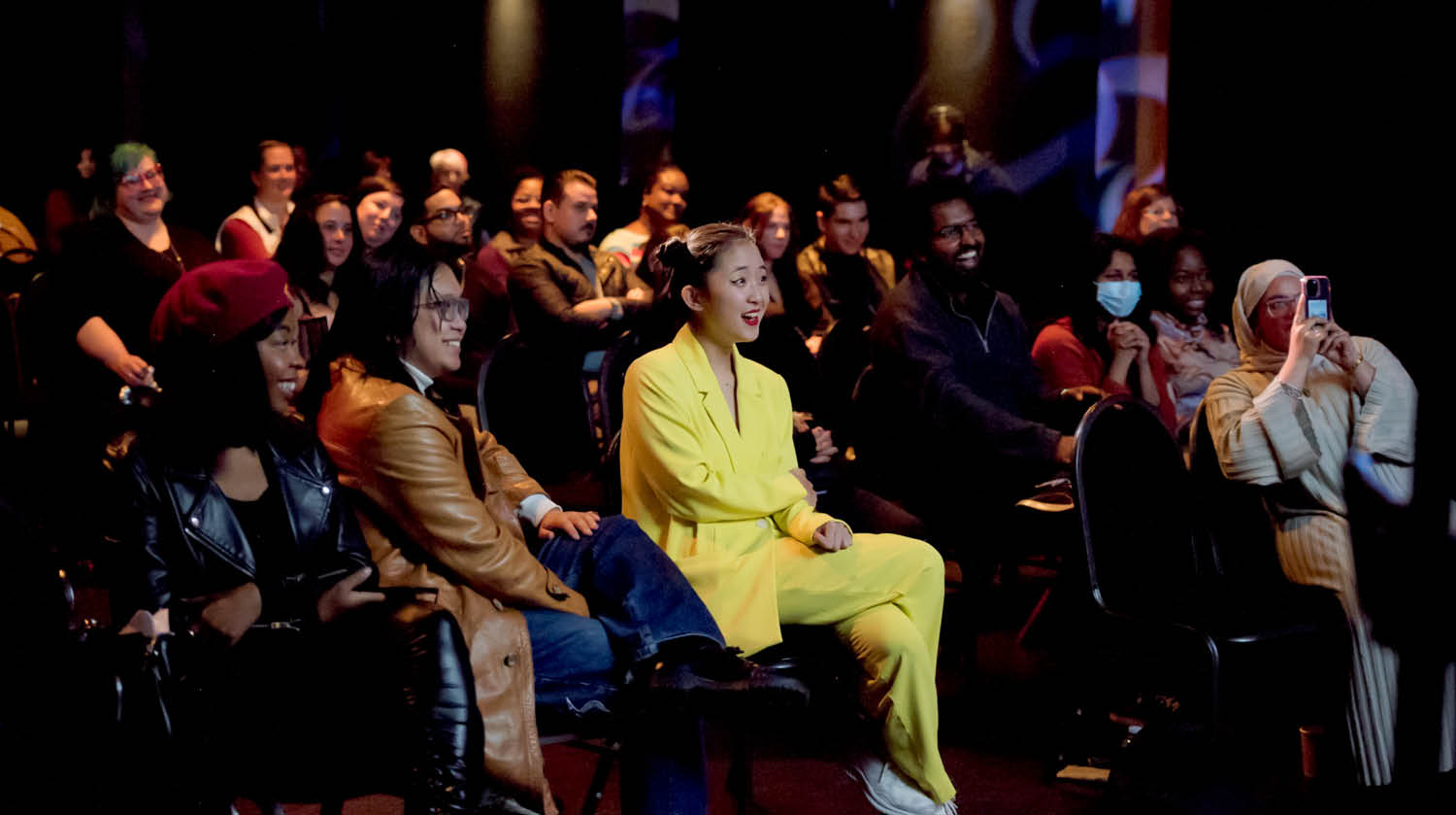 Image of audience members smiling and enjoying a performance in The Rose Studio. One woman holds her phone in front of her to capture the show. Another woman in a yellow matching suit smiles enthusiastically as she observes the performance. 