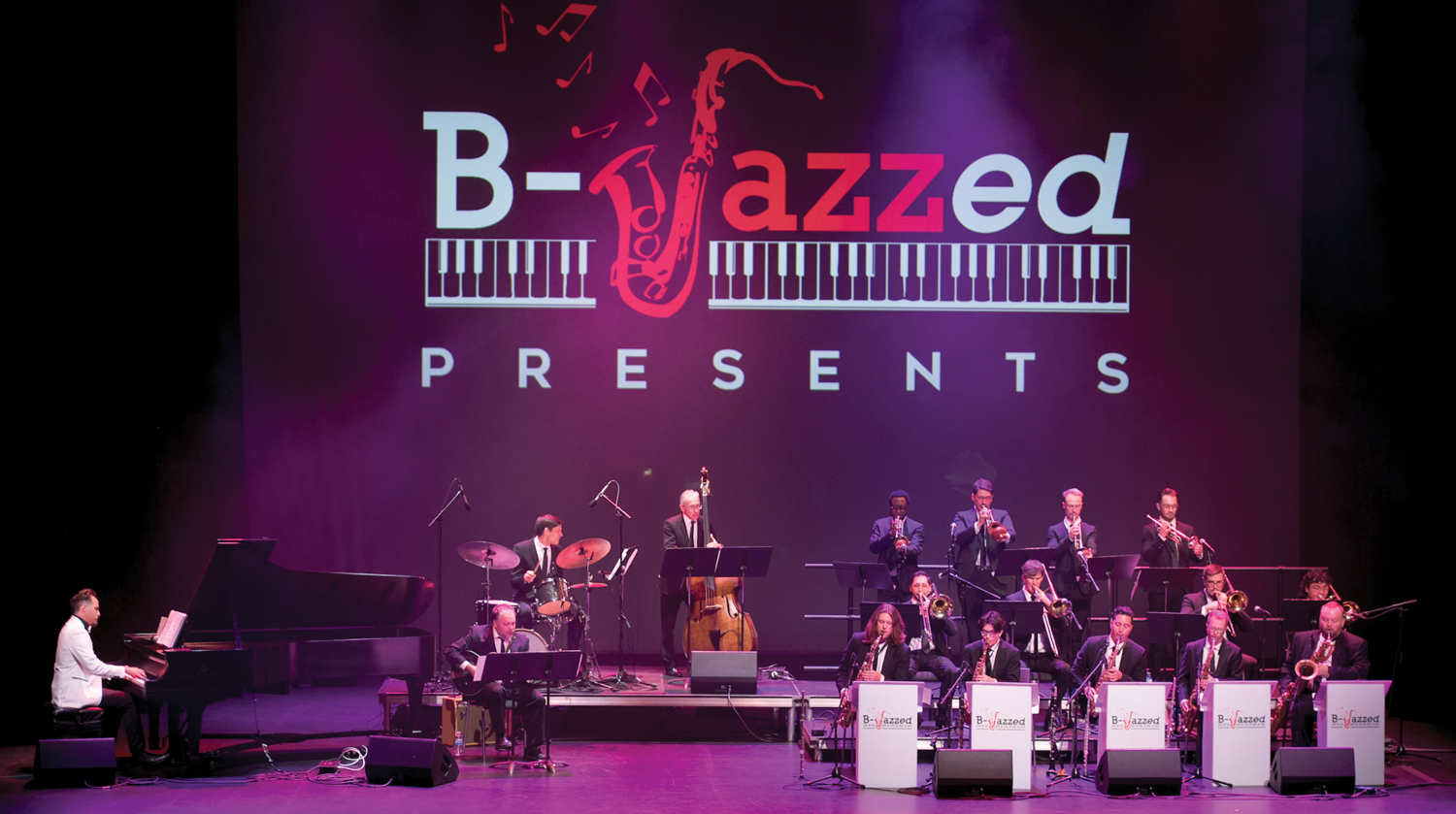 Image of the B-Jazzed band performing on The Rose Mainstage. To the left of the image, several musicians play an array of instruments, including the saxophone, trumpet, and drums. To the right of the image, artist Carmen Spada plays the piano. Behind the performs, a projection reads "B-Jazzed Presents."